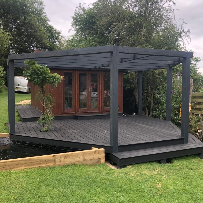 Wooden cabin with a dark pergola and spacious deck, surrounded by garden and pond.