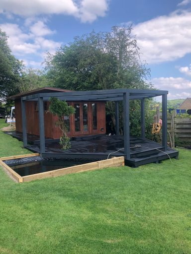 A wooden garden cabin with a black pergola with decking, surrounding garden next to a pond.